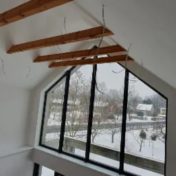 Poddasze z drewnianymi belkami stropowymi i dużym oknem z widokiem na zimowy krajobraz.