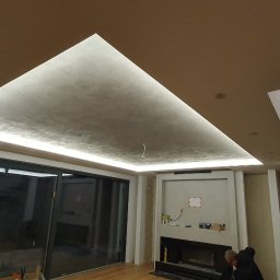 BUDAR - Nowoczesne wnętrze z podwieszanym sufitem oświetlonym taśmą LED, ściany częściowo wykończone gładzią, widoczne schody z surowego betonu, duże przesuwne okno tarasowe.