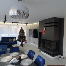 BUDAR - Panoramiczny widok na salon z kominkiem obłożonym szarą okładziną, świąteczną choinką, niebieską sofą i stołem z nakryciem, oświetlony nowoczesną lampą wiszącą z kryształkami.