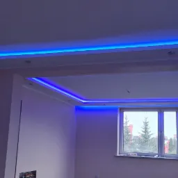 Wykończone wnętrze z podwieszanym sufitem, oświetleniem LED w kolorze niebieskim, widok na okno z drzewami na zewnątrz.