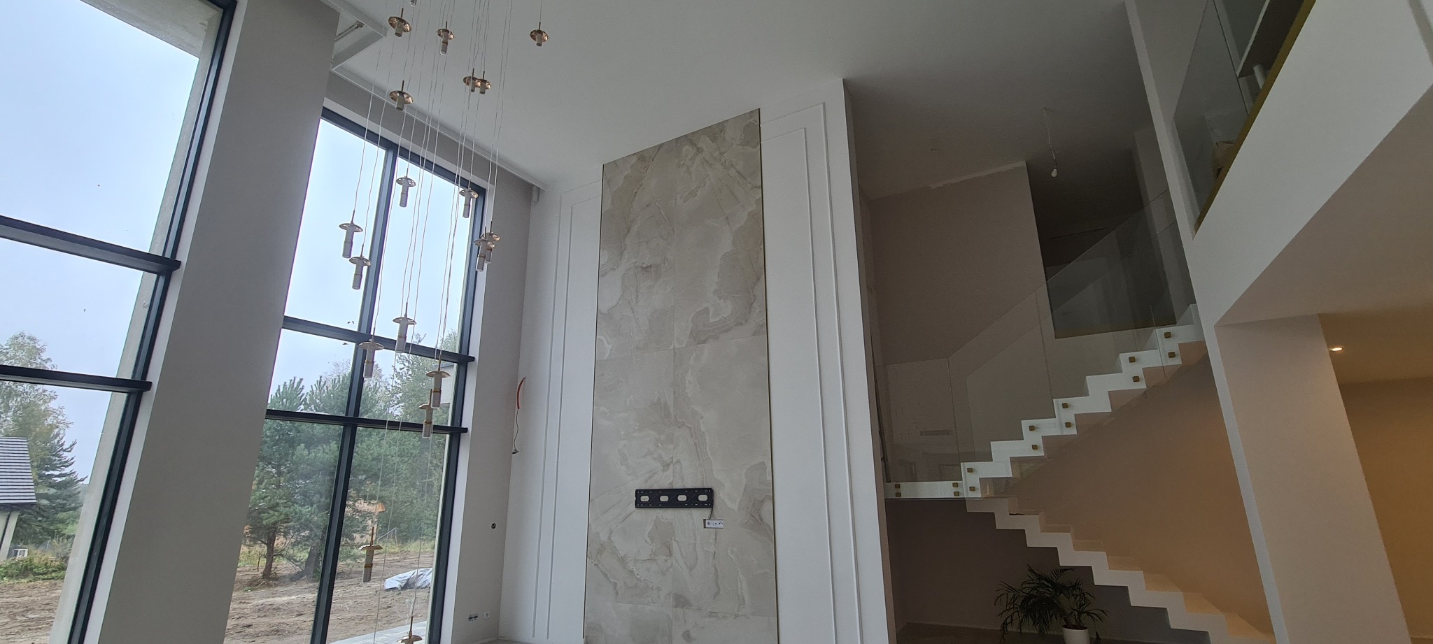 Wnętrze domu w trakcie wykańczania, widoczne duże okno, ściana wyłożona płytami imitującymi marmur, nowoczesne schody z przeszkloną balustradą oraz wisząca lampa.