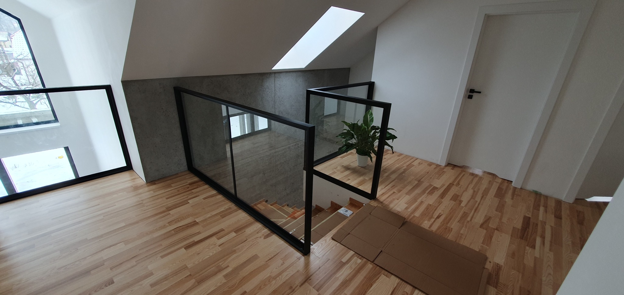 Szklana balustrada z czarną ramą na schodach z drewnianą podłogą, widok z góry, roślina doniczkowa za balustradą, zabezpieczone schody kartonem.