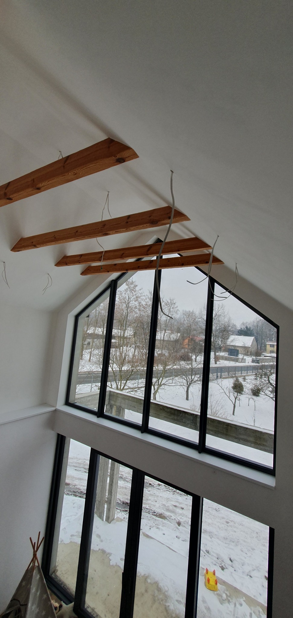 Poddasze z drewnianymi belkami stropowymi i dużym oknem z widokiem na zimowy krajobraz.