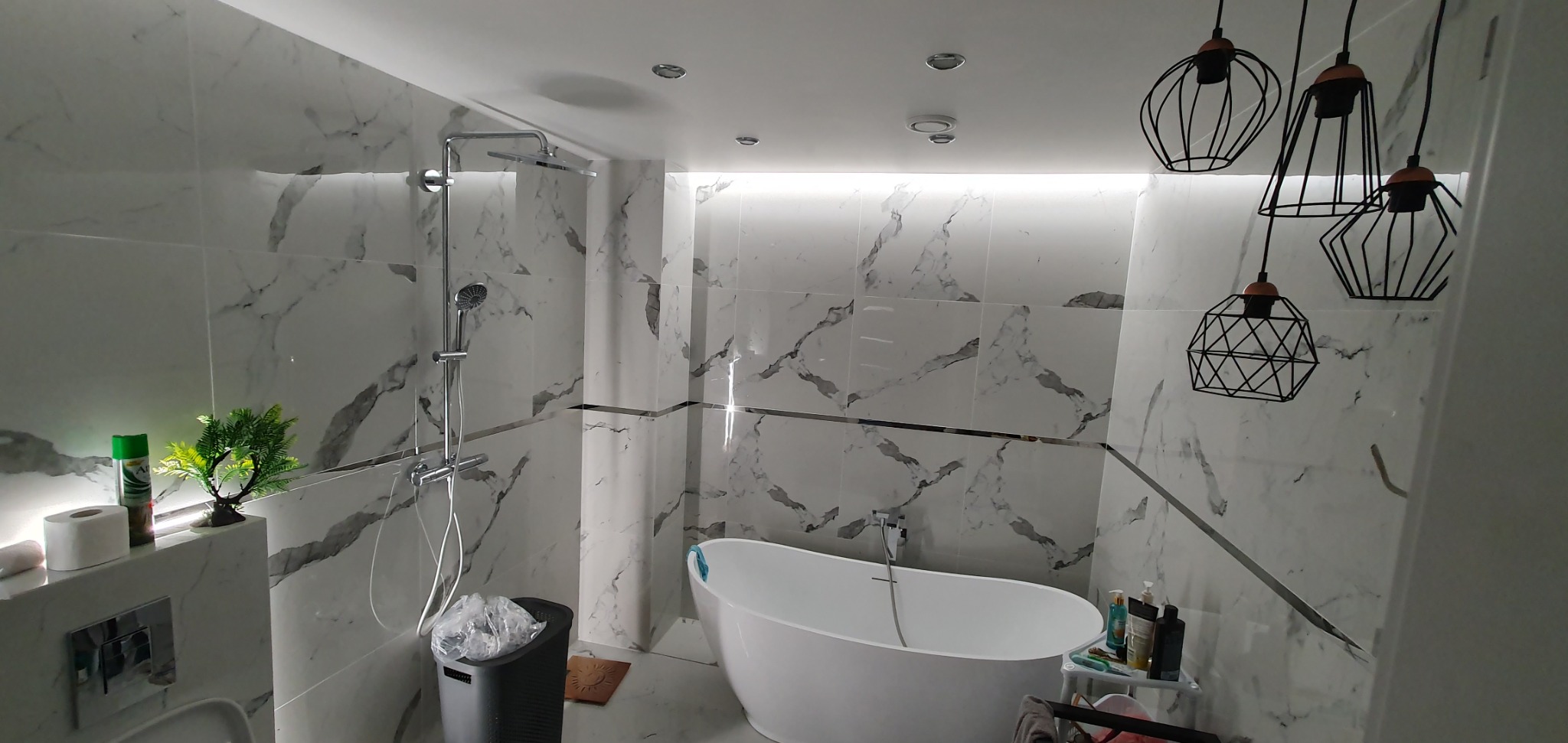 Nowoczesna łazienka z białą wanną wolnostojącą, chromowaną armaturą, marmurowymi płytkami na ścianach i geometrycznymi lampami wiszącymi.