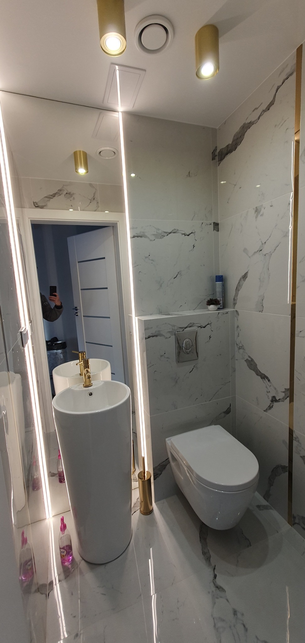 Nowoczesna łazienka z marmurowymi płytkami, wolnostojącą umywalką z złotą armaturą, podświetlanym lustrem i wiszącą toaletą.