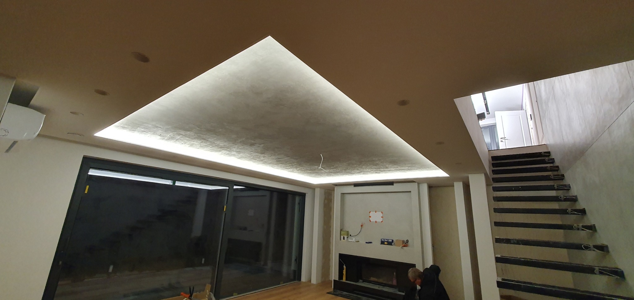 Nowoczesne wnętrze z podwieszanym sufitem oświetlonym taśmą LED, ściany częściowo wykończone gładzią, widoczne schody z surowego betonu, duże przesuwne okno tarasowe.