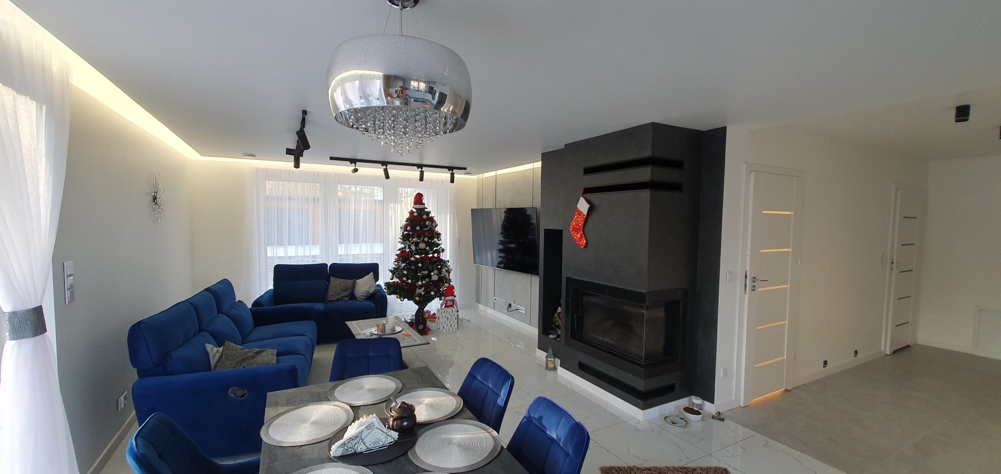 Panoramiczny widok na salon z kominkiem obłożonym szarą okładziną, świąteczną choinką, niebieską sofą i stołem z nakryciem, oświetlony nowoczesną lampą wiszącą z kryształkami.
