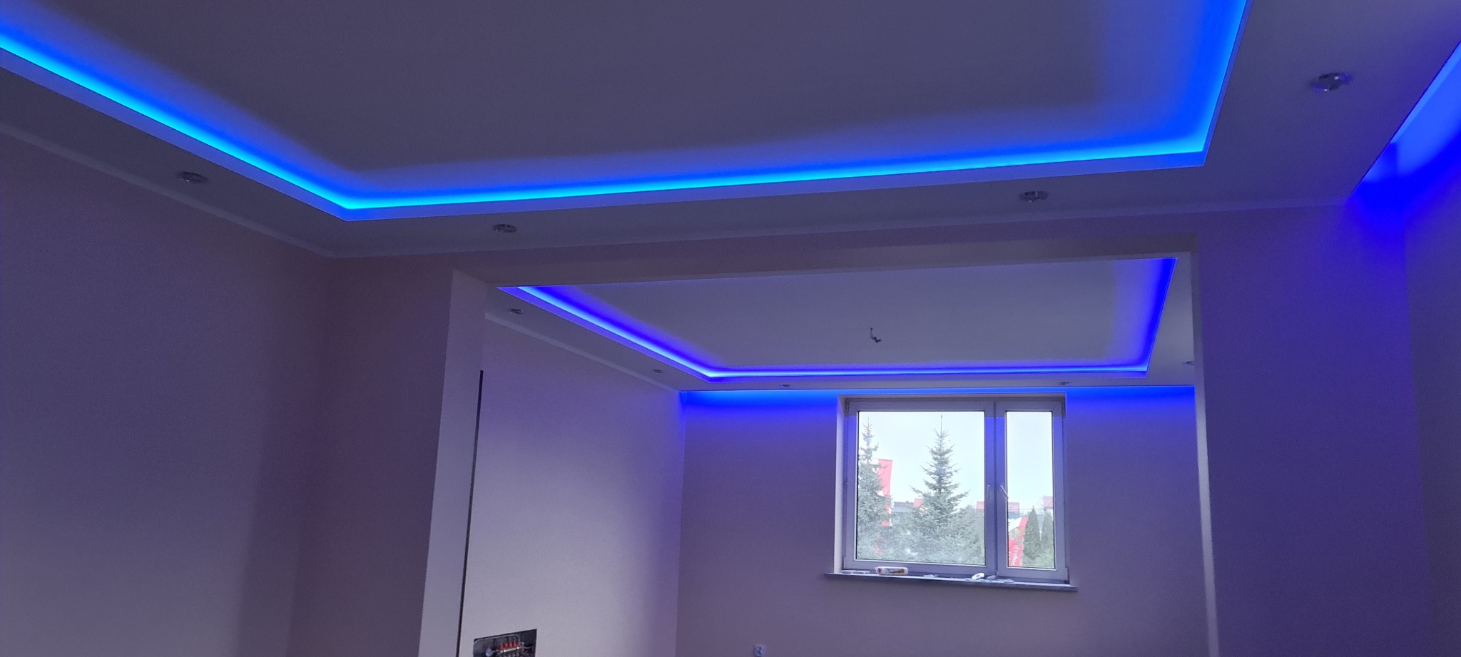 Wykończone wnętrze z podwieszanym sufitem, oświetleniem LED w kolorze niebieskim, widok na okno z drzewami na zewnątrz.