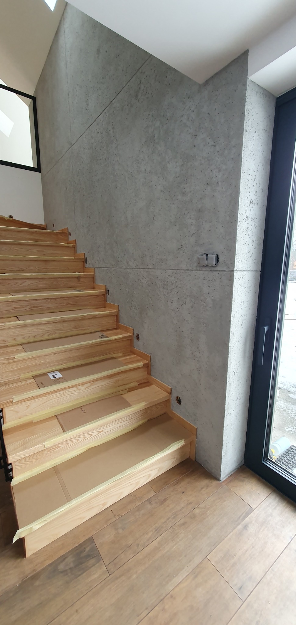 Surowe drewniane schody zabezpieczone kartonem i taśmą malarską, prowadzące w górę, na tle ściany wykończonej szarą, betonową fakturą i drewnianej podłogi.