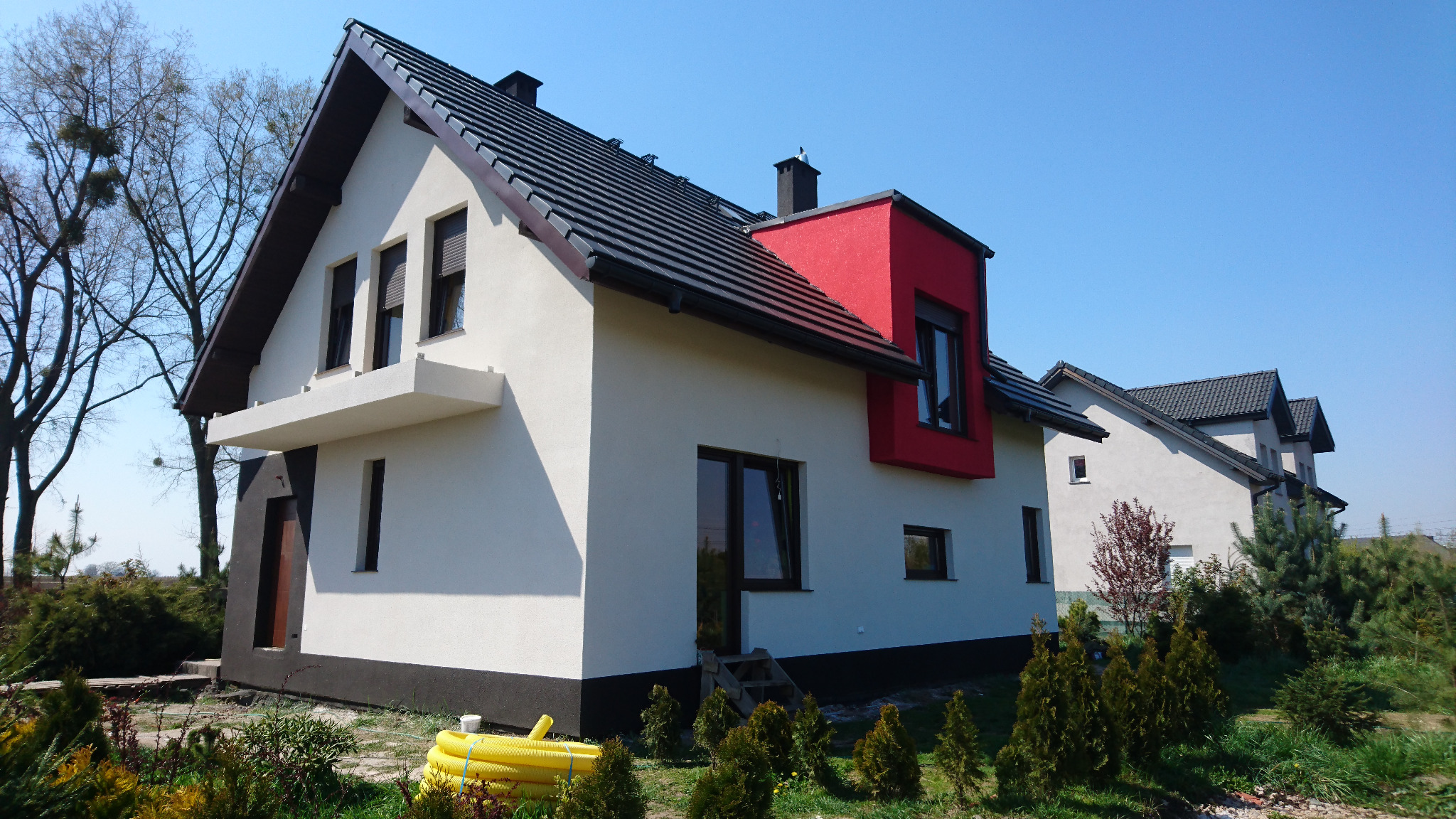 Nowoczesny dom jednorodzinny z białą elewacją, czarnym dachem i czerwonym akcentem na lukarnie, otoczony zielenią.