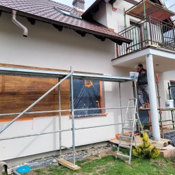 P.H.U Len-Bud - Pracownik maluje elewację domu jednorodzinnego ze schodami i balkonem, widoczne rusztowania, fragment drewnianej okładziny elewacyjnej oraz zabezpieczone okno folią.