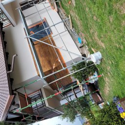 P.H.U Len-Bud - Dom w trakcie prac elewacyjnych, widoczne rusztowania, częściowo otynkowana ściana, zabezpieczone okna folią, wiadro z zaprawą i fragment ogrodu z kwiatami.