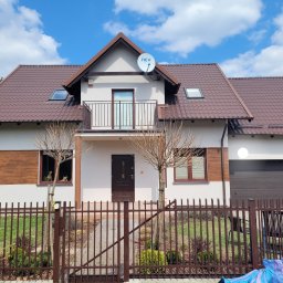 P.H.U Len-Bud - Dwupiętrowy dom z brązowym dachem i balkonem, widoczny przez metalowe ogrodzenie; na elewacji zamontowana antena satelitarna.