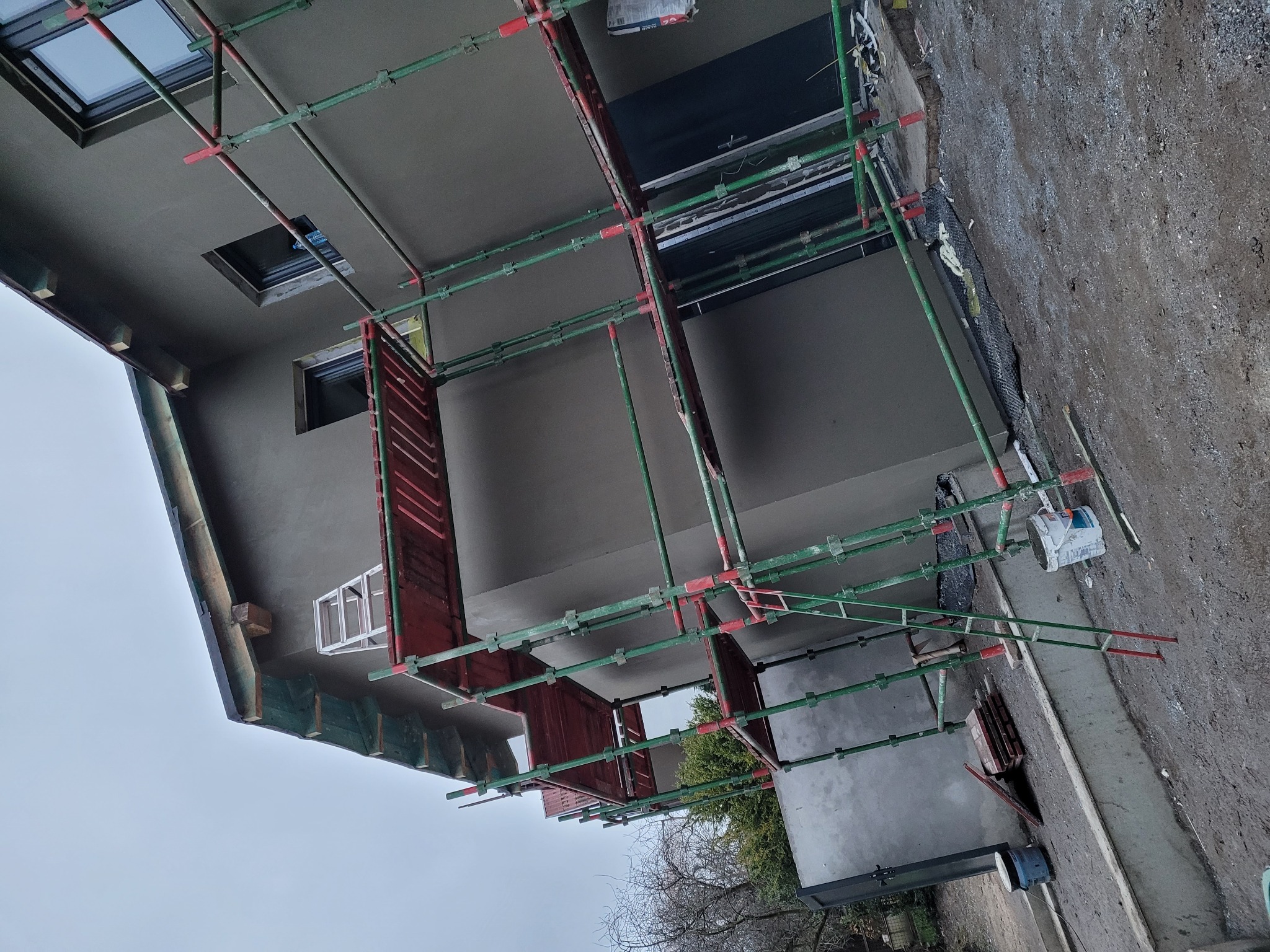 Świeżo otynkowana elewacja domu jednorodzinnego z widocznymi oknami dachowymi, częściowo zasłonięta rusztowaniem, z elementami izolacji fundamentów przy gruncie.