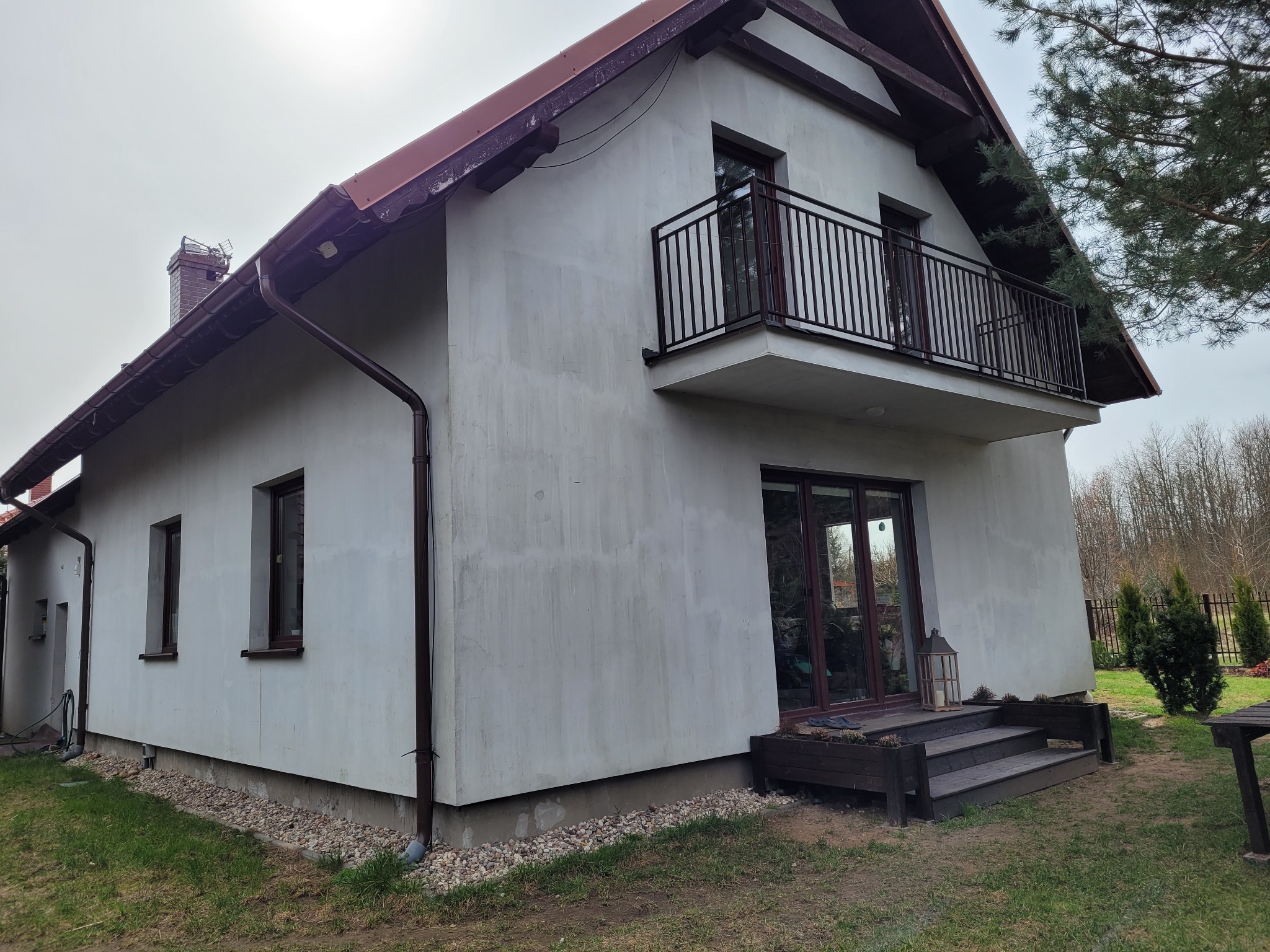 Dwupiętrowy dom z balkonem i widocznymi śladami po wcześniejszych pracach na elewacji, brązowe rynny i stolarka okienna, otoczenie trawnik i drzewa.
