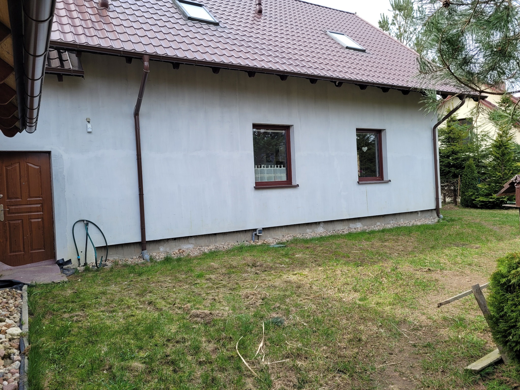 Ściana domu jednorodzinnego po tynkowaniu, z widocznymi oknami w brązowych ramach, rynną spustową i fragmentem trawnika z pozostałościami po koszeniu.