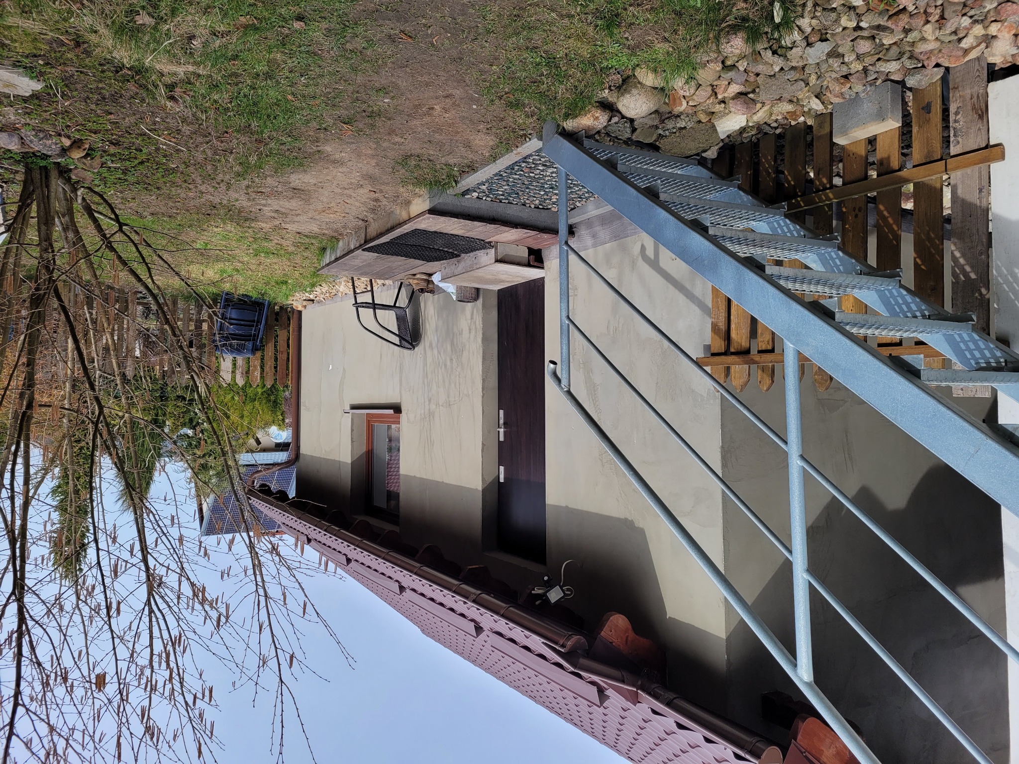 Elewacja domu z zewnętrznymi metalowymi schodami, widoczne tynkowanie i brązowe okno oraz drzwi, dach pokryty dachówką, w tle trawa i drzewa.