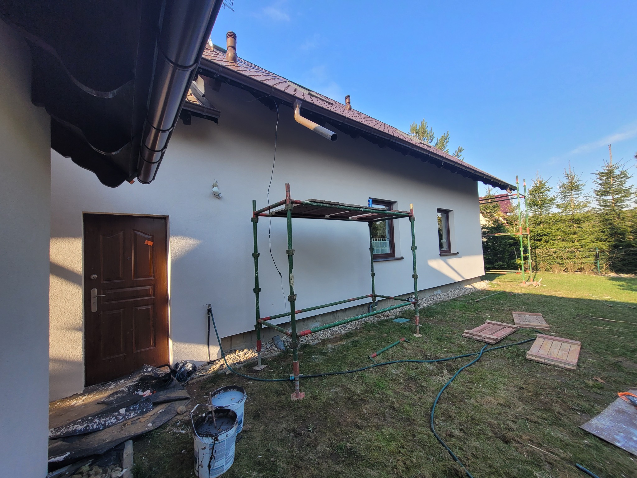 Dom jednorodzinny w Pszczółkach z jasną elewacją w trakcie prac wykończeniowych; widoczne rusztowanie, wiaderka z farbą, zabezpieczenia przy drzwiach wejściowych i przygotowane płyty chodnikowe...