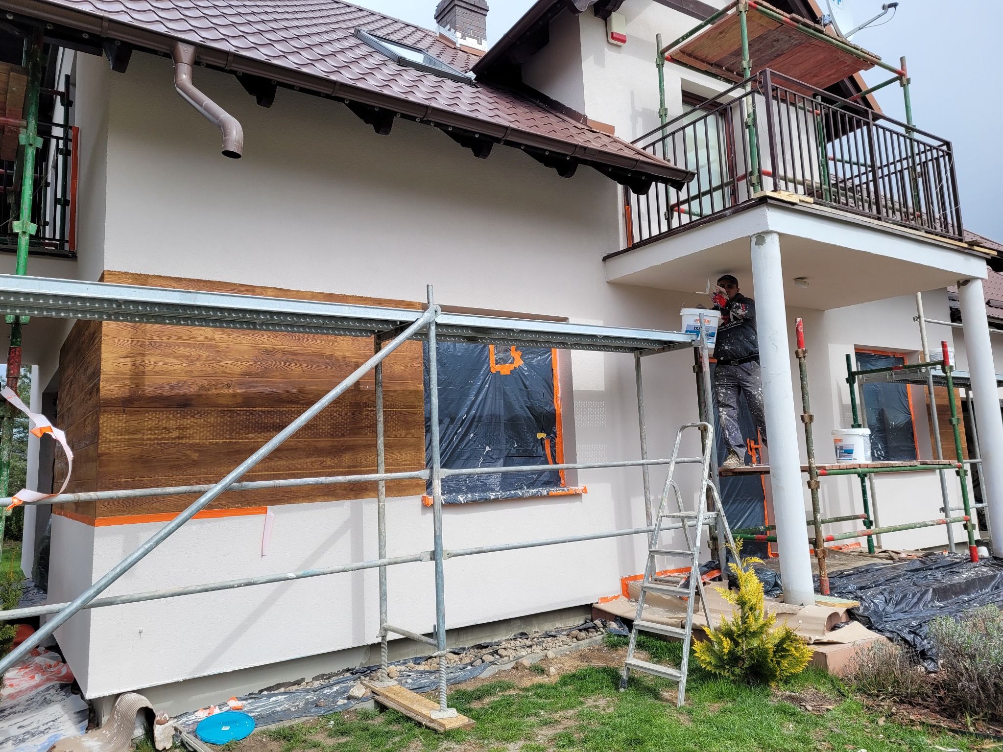 Pracownik maluje elewację domu jednorodzinnego ze schodami i balkonem, widoczne rusztowania, fragment drewnianej okładziny elewacyjnej oraz zabezpieczone okno folią.