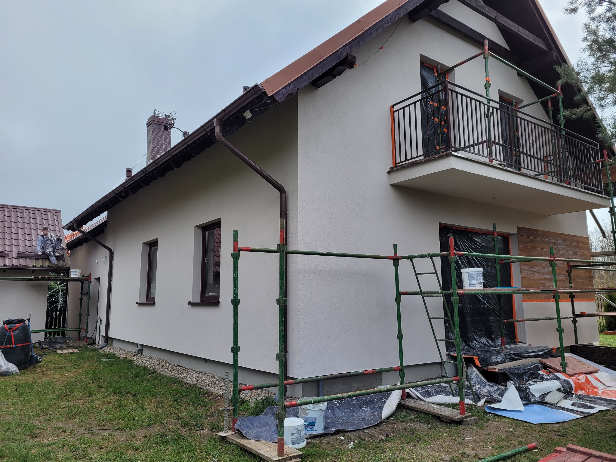 Dwukondygnacyjny dom z beżową elewacją w trakcie prac wykończeniowych, rusztowania przy balkonie i wejściu, widoczne wiaderka z farbą i zabezpieczenia okien folią.
