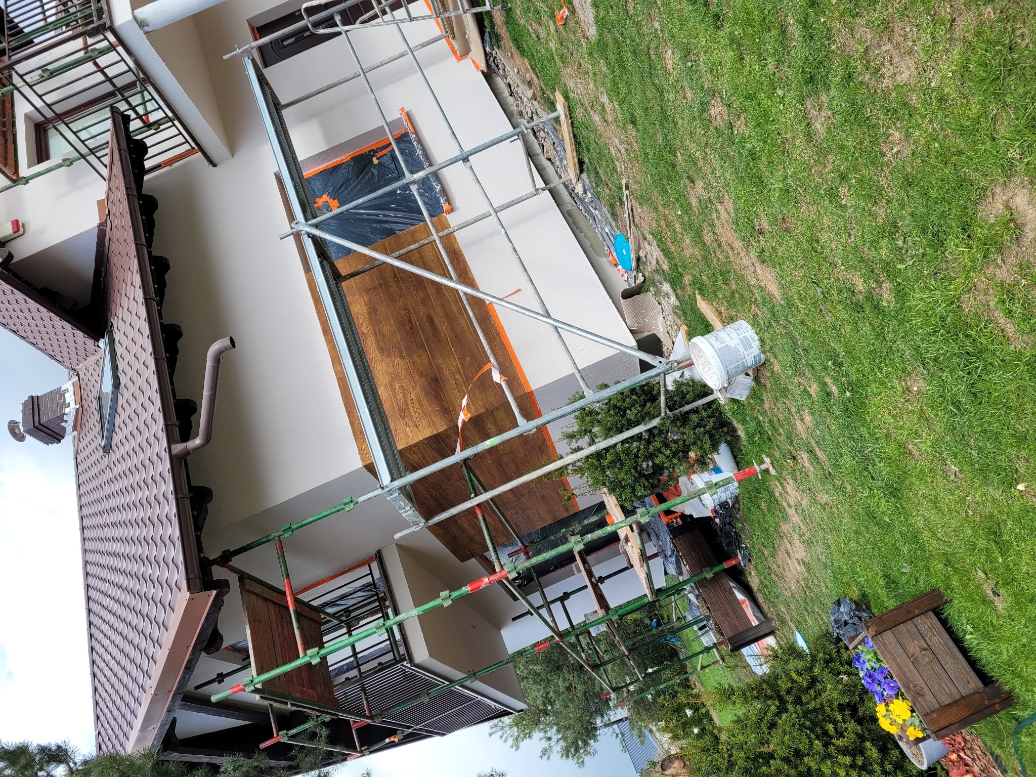 Dom w trakcie prac elewacyjnych, widoczne rusztowania, częściowo otynkowana ściana, zabezpieczone okna folią, wiadro z zaprawą i fragment ogrodu z kwiatami.