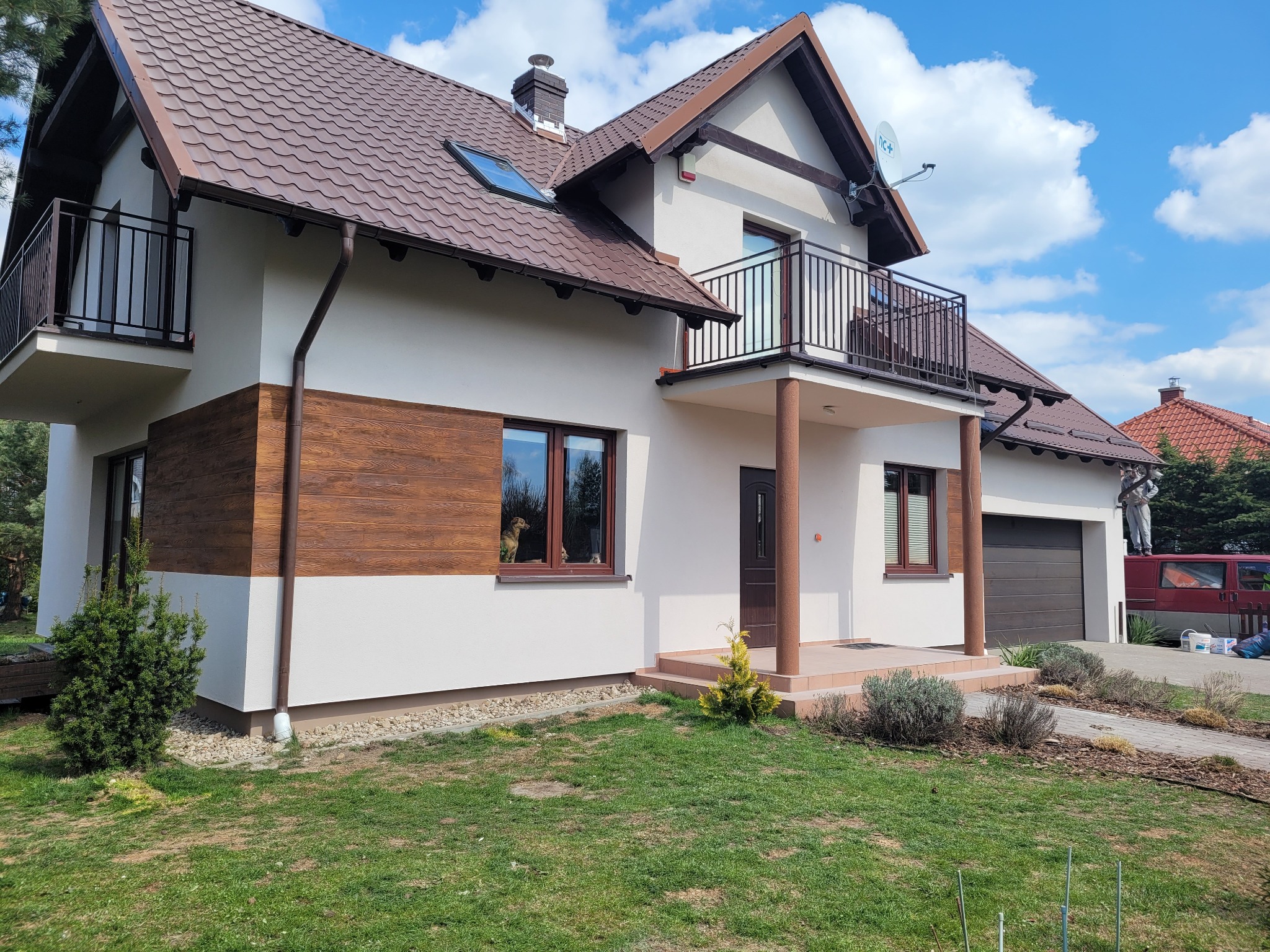 Dwukondygnacyjny dom z brązowym dachem, białymi ścianami i drewnianą elewacją, balkon z czarną balustradą, widok z ogrodu w słoneczny dzień.