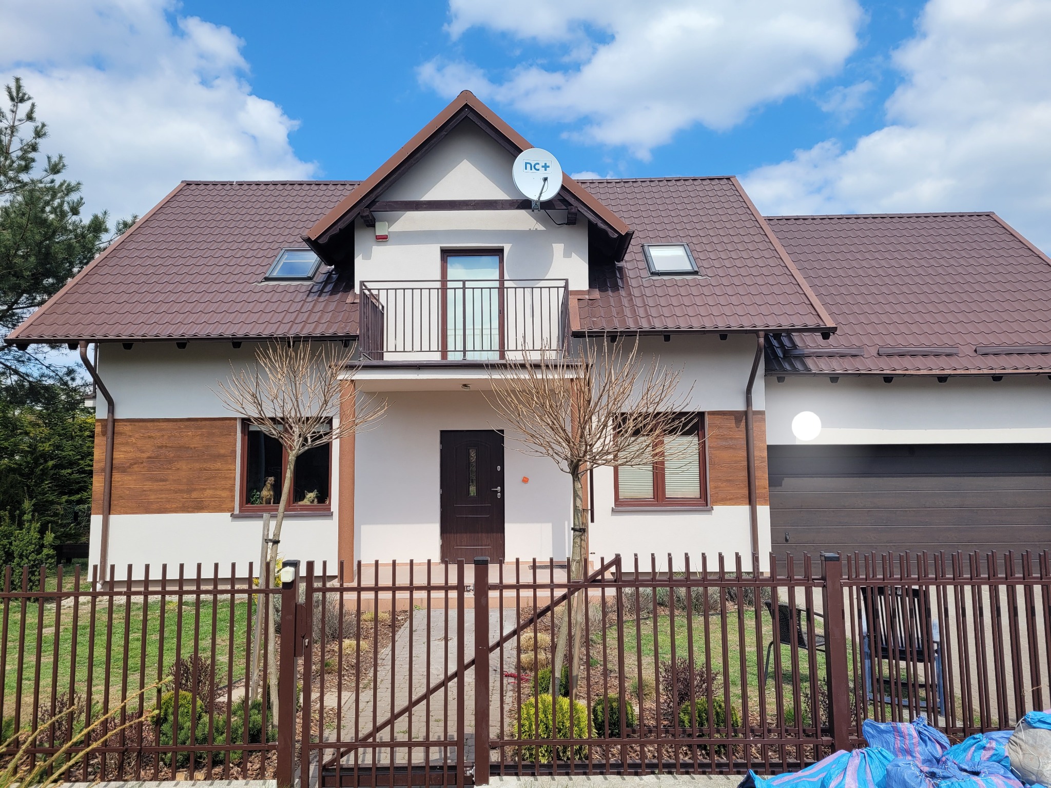 Dwupiętrowy dom z brązowym dachem i balkonem, widoczny przez metalowe ogrodzenie; na elewacji zamontowana antena satelitarna.