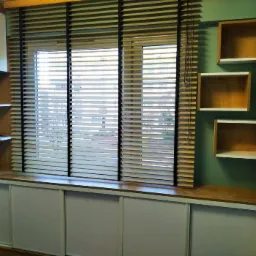 Okno z drewnianymi żaluzjami, szafka pod oknem z przesuwnymi drzwiami i wiszące drewniane półki w kształcie kostek, na ścianie w kolorze morskim.