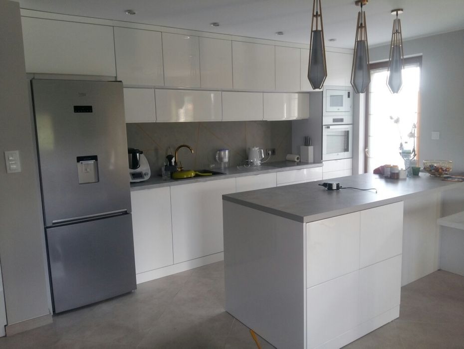 Nowoczesna, biała kuchnia z wyspą i szarym blatem, oświetlona trzema wiszącymi lampami w geometrycznym stylu, z widoczną srebrną lodówką i elementami wyposażenia.