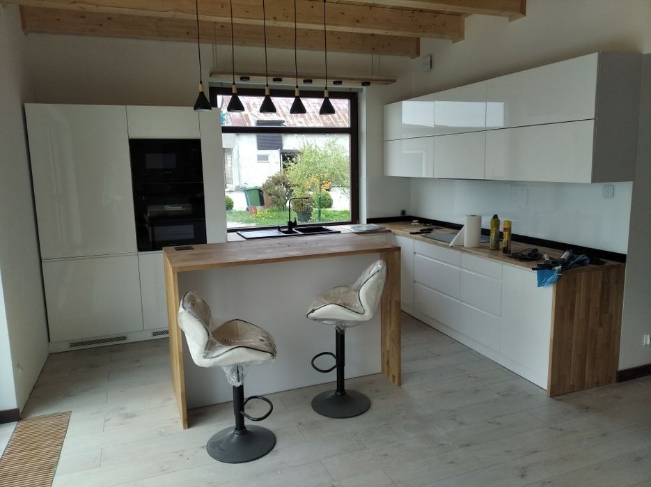 Nowoczesna, jasna kuchnia z białymi szafkami na wysoki połysk, drewnianym blatem i barkiem, oraz dwoma krzesłami barowymi jeszcze w folii ochronnej. Widoczne drewniane belki stropowe i lampy...