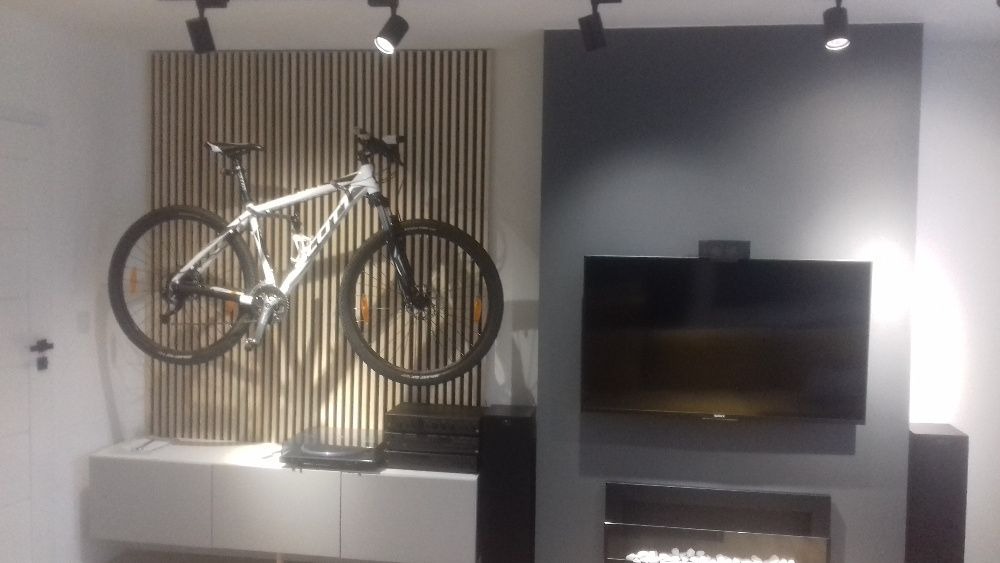 Biały rower górski zawieszony na ścianie z drewnianych listew w nowoczesnym salonie z szarą ścianą, telewizorem i kominkiem elektrycznym.