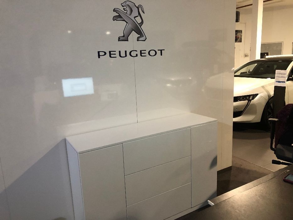 Biała komoda z trzema szufladami i drzwiczkami, umieszczona pod logo Peugeot w salonie samochodowym, z widocznym fragmentem białego samochodu w tle.