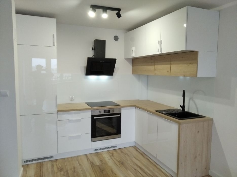 Nowoczesna kuchnia z białymi lakierowanymi frontami, drewnopodobnym blatem i czarną armaturą, oświetlona trzypunktową lampą sufitową.