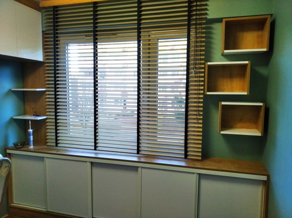 Okno z drewnianymi żaluzjami, szafka pod oknem z przesuwnymi drzwiami i wiszące drewniane półki w kształcie kostek, na ścianie w kolorze morskim.