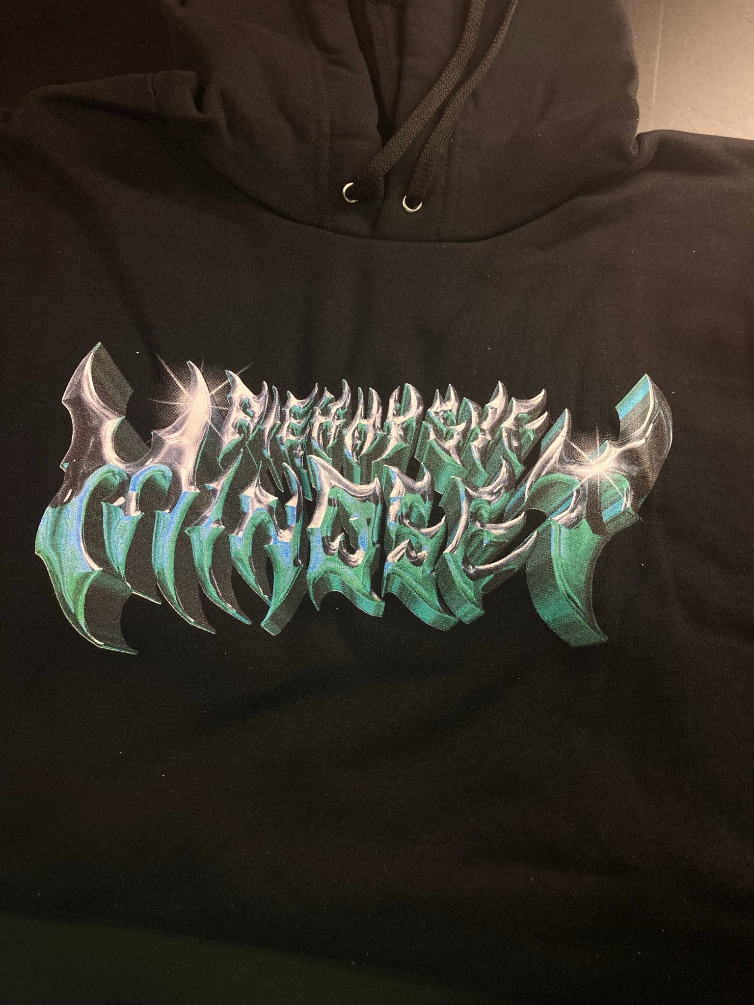Czarna bluza z kapturem, na której znajduje się duży, metaliczny nadruk w odcieniach zieleni i srebra, przedstawiający abstrakcyjny napis o ostrych krawędziach.