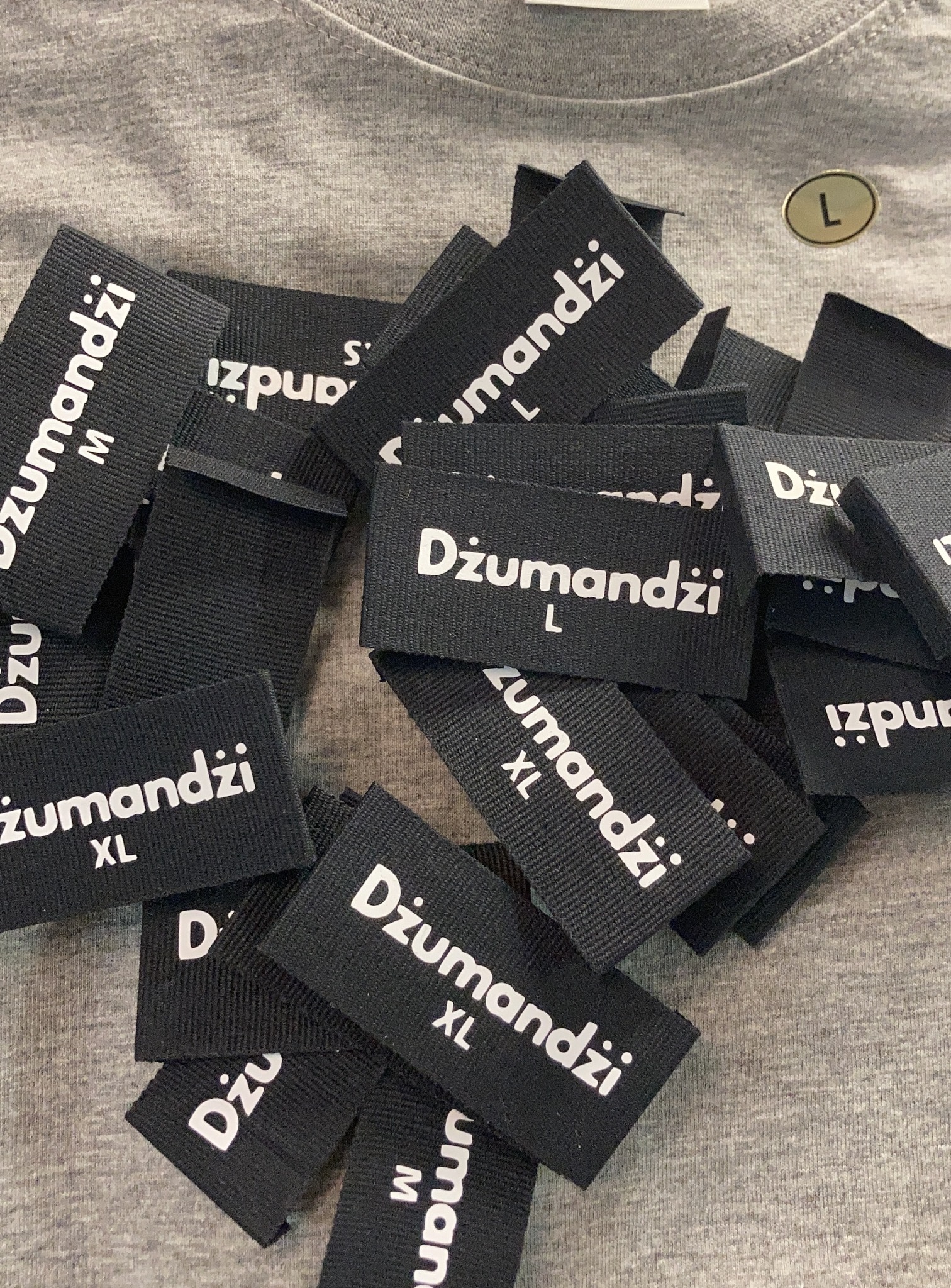 Stos czarnych metek odzieżowych z białym nadrukiem nazwy firmy 'Dżumandzi' i rozmiarów M, L, XL, leżących na szarej koszulce.