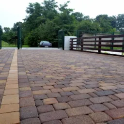 Ułożona, wielobarwna kostka brukowa przed posesją z drewnianym ogrodzeniem i metalową bramą w tle.