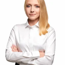 Portret kobiety w białej koszuli z delikatnym wzorem i skrzyżowanymi ramionami na białym tle.