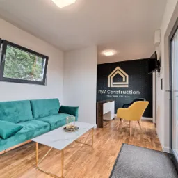 Wnętrze nowocześnie urządzonego salonu w domu z zieloną sofą, marmurowym stolikiem kawowym na złotym stelażu oraz żółtym krzesłem na tle czarnej ściany z białym logo firmy RW Construction.