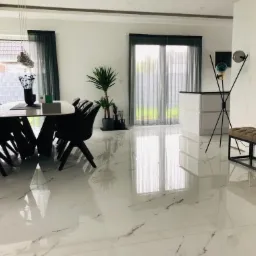 Jasne, przestronne wnętrze jadalni z marmurową podłogą, nowoczesnym stołem otoczonym czarnymi krzesłami, wysoką lampą z kolorowymi kulami i minimalistyczną ławką w tle.