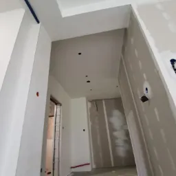 Wnętrze w trakcie prac wykończeniowych, widoczne ściany przygotowane do malowania, z widocznymi otworami na gniazdka elektryczne i oświetlenie, częściowo pokryte masą szpachlową.