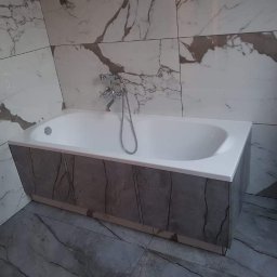 SZWAGRO-BUD - Biała wanna obudowana panelami imitującymi marmur, z chromowaną armaturą, na tle ściany wyłożonej płytkami z wzorem marmuru.