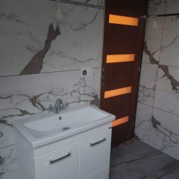 SZWAGRO-BUD - Umywalka z szafką w łazience wykończonej płytkami imitującymi marmur, obok drewniane drzwi z poziomymi przeszkleniami.