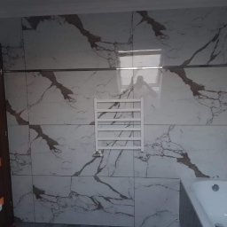 SZWAGRO-BUD - Wykończona łazienka z dużymi, białymi płytkami imitującymi marmur z brązowymi żyłami, grzejnik drabinkowy na ścianie, widoczna wanna i fragment drzwi z pomarańczowymi wstawkami.