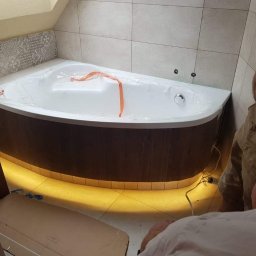 SZWAGRO-BUD - Narożna wanna z hydromasażem w folii ochronnej, podświetlona od dołu, częściowo widoczni dwaj instalatorzy w trakcie montażu, płytki ceramiczne na ścianach i podłodze.