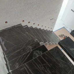 Schody wyłożone czarnym marmurem z widocznymi białymi żyłami, zabezpieczone folią ochronną podczas prac wykończeniowych, z widocznymi puszkami elektrycznymi w ścianie.