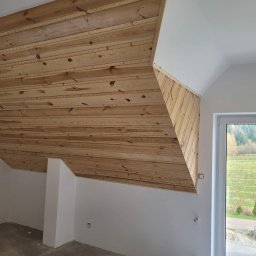 SZWAGRO-BUD - Drewniana boazeria na poddaszu, widok na świeżo pomalowane ściany i okno z widokiem na zewnątrz.