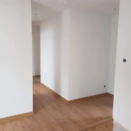 SZWAGRO-BUD - Świeżo pomalowane na biało wnętrze z drewnianą podłogą, widoczne wystające przewody elektryczne i puste gniazdka.