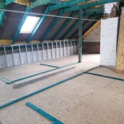 SZWAGRO-BUD - Poddasze w trakcie adaptacji: widoczne ocieplenie wełną mineralną, metalowy stelaż pod płyty gipsowo-kartonowe, surowe drewniane belki konstrukcyjne i częściowo ułożona podłoga z płyt OSB.