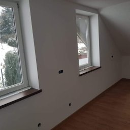 SZWAGRO-BUD - Poddasze po malowaniu na biało, widoczne dwa okna z ciemnymi parapetami, brązowe panele podłogowe, puszki elektryczne na ścianach czekające na montaż osprzętu.